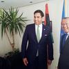 رجلان يرتديان بدلات رسمية يقفان مبتسمين لالتقاط صورة في الداخل، مع وجود أعلام في الخلفية.