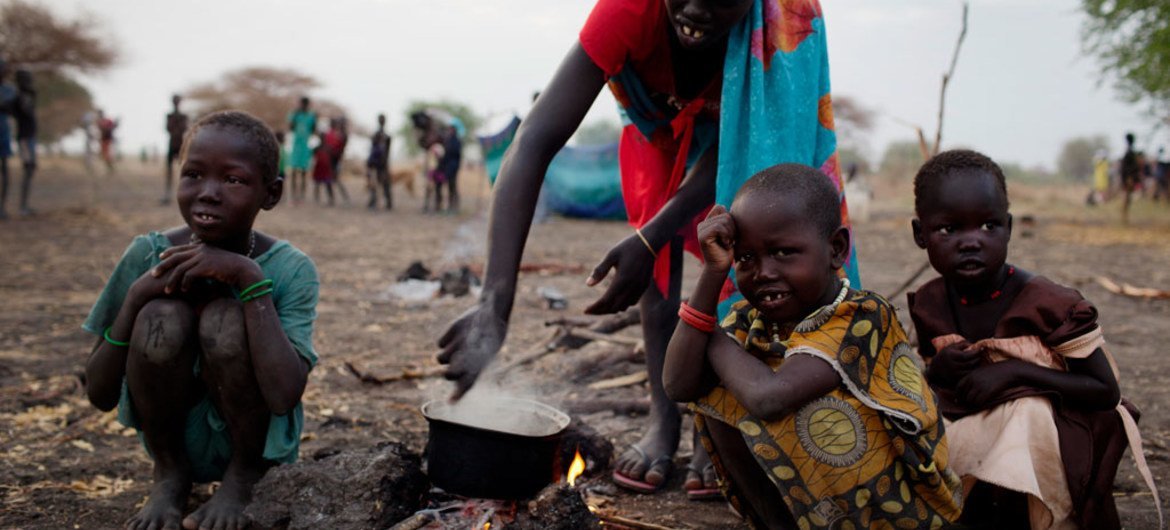 A woman cooks a meal outside in the open air with her three children where she is camping with her family the night before being able to register for a ration card  in Thanyang, South Sudan.