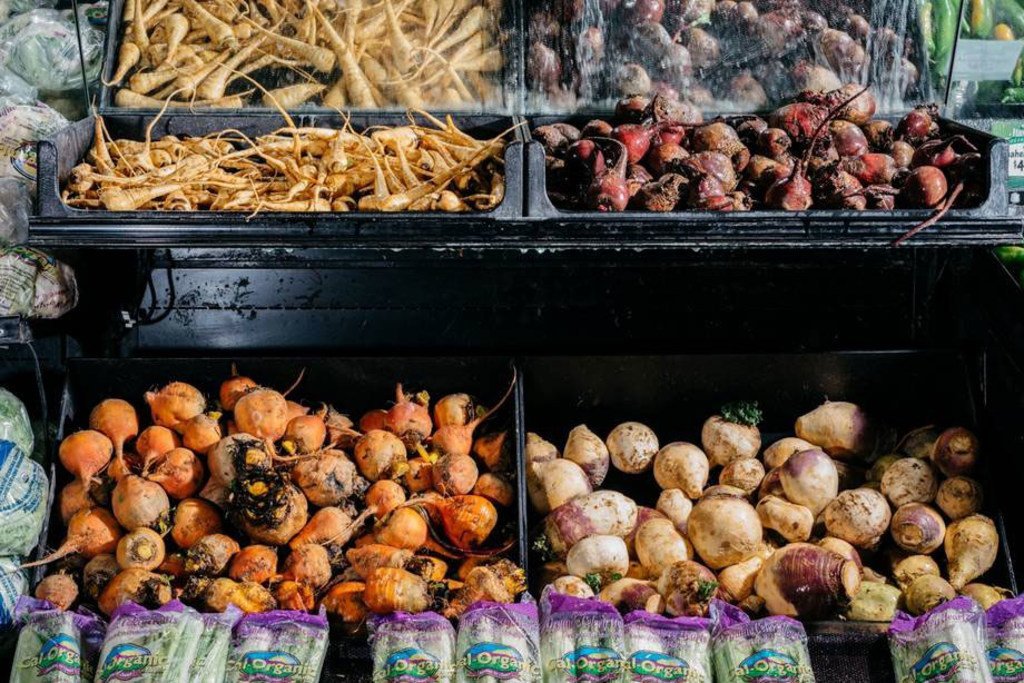 Fresh root vegetables, including parsnips, turnips, and beets, are displayed in bins at a market. Bags of Cal Organic greens are in the foreground.