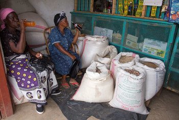 Two women sit in a shop in Kenya, drinking from a bottle and a plastic cup, surrounded by large sacks of sugar.