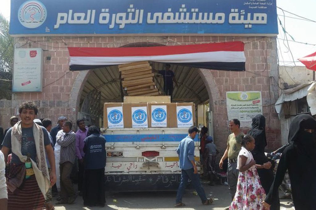A truck loaded with WHO-branded boxes unloads supplies at the Al-Thawra Hospital in Yemen, with a crowd of people gathered nearby.
