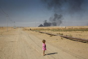 A young internally displaced Iraqi girl stands on a dusty path at the edge of Debaga camp, near Mosul in northern Iraq. Smoke from oil fires can be seen in the background.