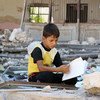 A boy, displaced from the ongoing conflict in Syria, sits and reads a book in the rubble of destroyed buildings.