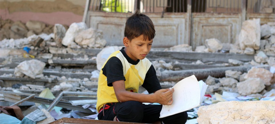 A boy, displaced from the ongoing conflict in Syria, sits and reads a book in the rubble of destroyed buildings.