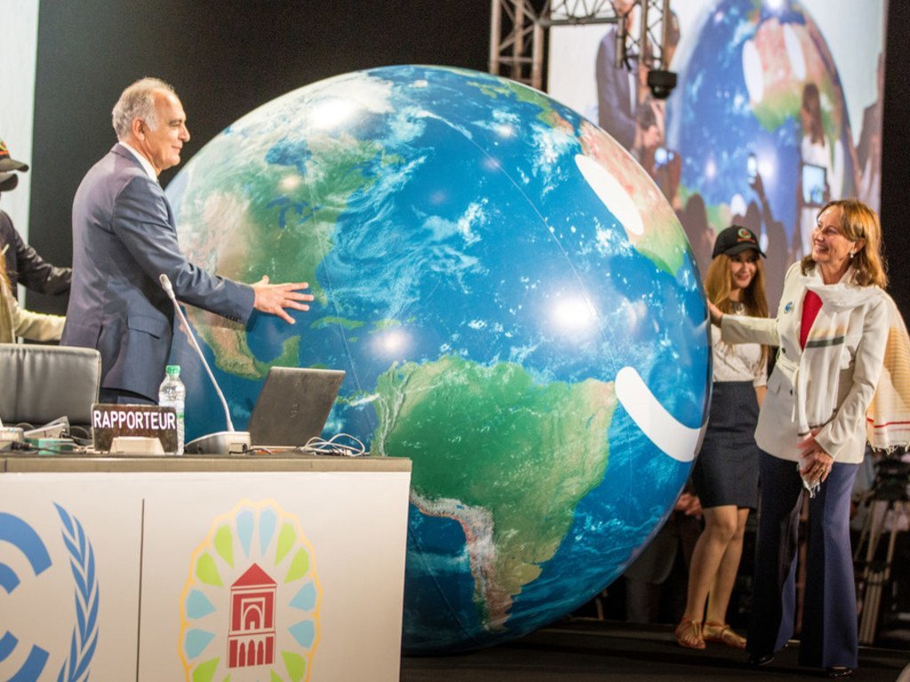 A man and a woman stand on a stage with a giant inflatable globe, next to a podium with a 'RAPPORTEUR' placard and the UNFCCC logo.