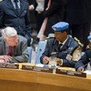 An older man in a suit leans in to speak with two uniformed officers wearing blue berets at a conference table with nameplates.