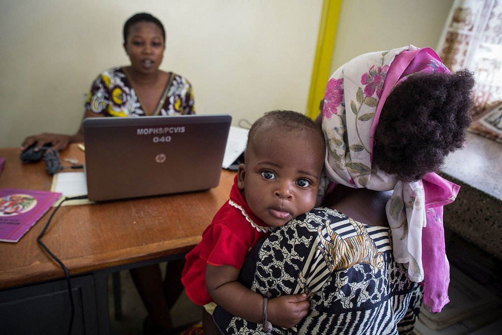 Mama akiwa amembeba mtoto wake huku mwanamke akifanya kazi kwenye kompyuta mpakato nyuma ya kliniki ya afya.