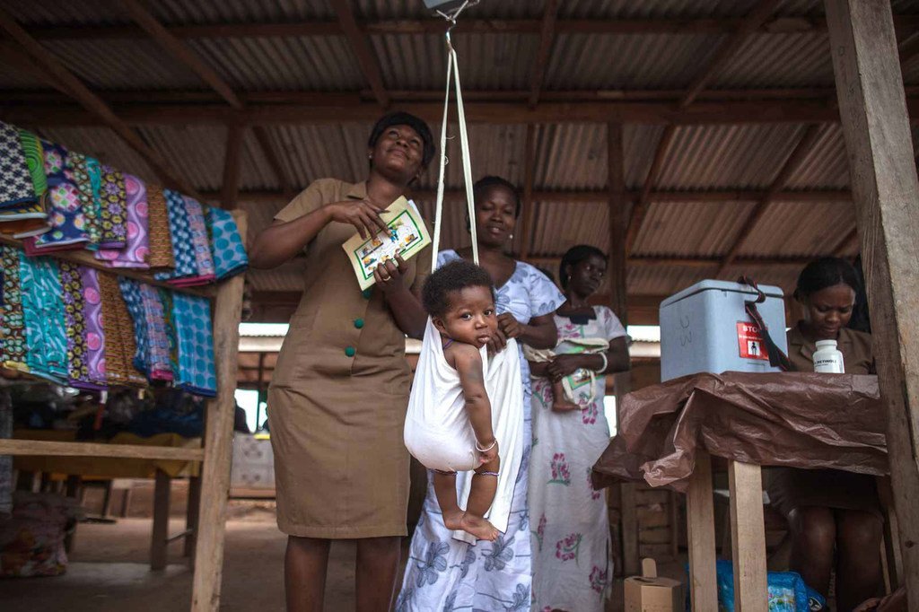 在加纳，一名社区卫生工作者在接种疫苗之前给一名婴儿称体重。 2012年，加纳成为非洲第一个同时引入肺炎球菌病和轮状病毒疫苗的国家，以应对世界上主要的两种致命儿童疾病：肺炎和腹泻。