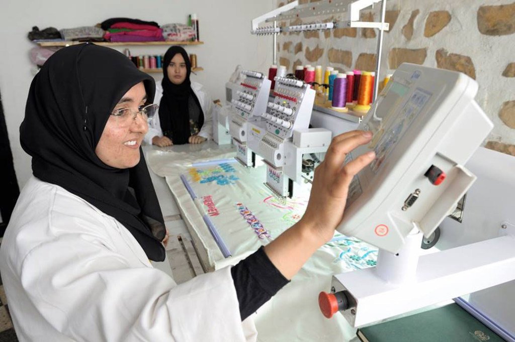 امرأة ترتدي معطف مختبر أبيض وحجابًا أسود تشغل آلة تطريز كبيرة في ورشة عمل، بينما تراقبها امرأة أخرى في الخلفية.