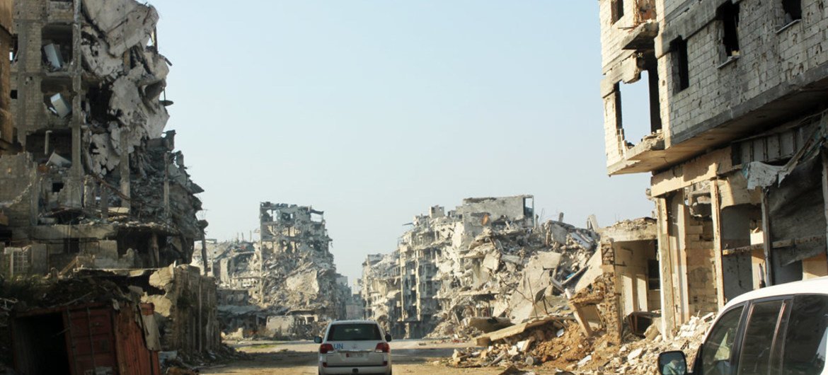 UN vehicles travel along a road lined with remnants of destroyed buildings, Homs, Syria. (file)