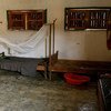 A room with simple wooden beds, one with a mosquito net, concrete floor, and windows with slatted shutters.