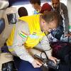 Un professionnel de santé en uniforme jaune et beige orné d'un écusson aux couleurs du drapeau kurde s'occupe d'un patient à l'intérieur d'une ambulance, vérifiant sa tension artérielle sous le regard d'autres personnes.