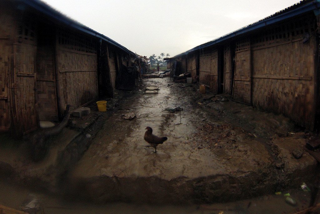 A flooded village in Myanmar with bamboo houses and a muddy path where a chicken is standing.