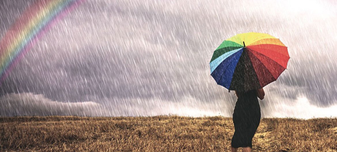 A woman with an umbrella walks in the rain in a field. To the left is a rainbow.