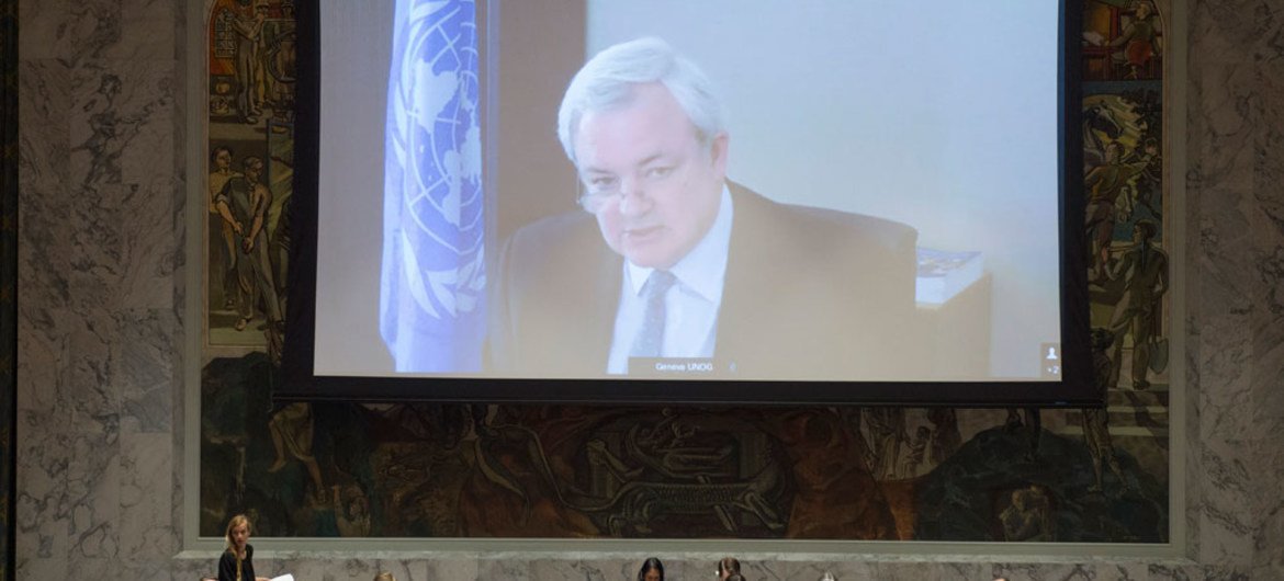 Lors d'une réunion d'information de ONU à Genève, un homme apparaît sur un grand écran, tandis que le public est assis autour d'une longue table dans une salle de conférence.