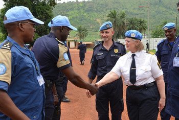A female UN peacekeeper in a white shirt and blue beret shakes hands with a male officer in a blue uniform during an outdoor meeting.