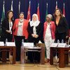 Five women stand at a formal conference table with nameplates, with the flags of Syria, Jordan, and Oman visible in the background.