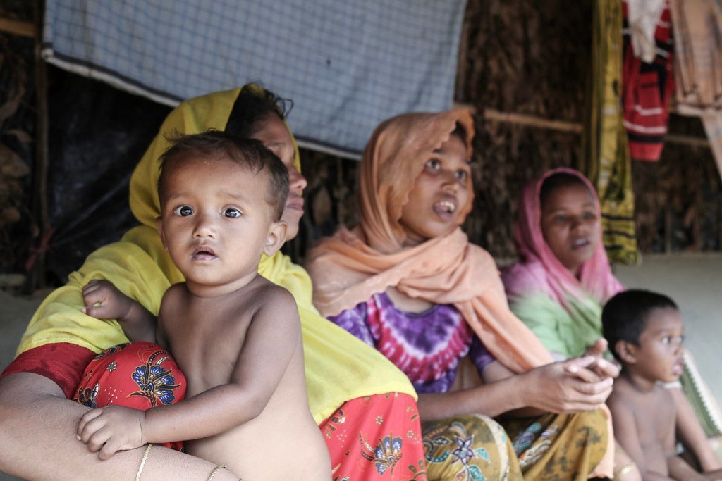 Une femme rohingya tient un bébé dans ses bras tandis que d'autres femmes et un jeune enfant les observent depuis un abri de fortune.