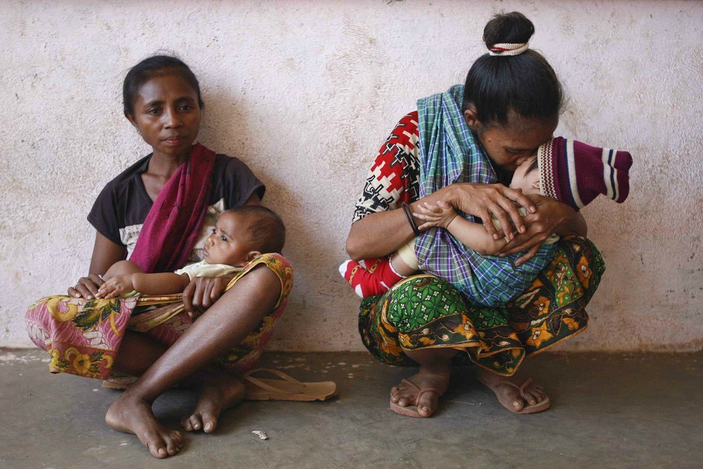 Duas mulheres estão agachadas contra uma parede, uma amamentando um bebê e a outra segurando um bebê no colo.