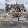 An orange excavator is clearing rubble from a destroyed building on a street in Mogadishu, Somalia, with two people standing nearby.