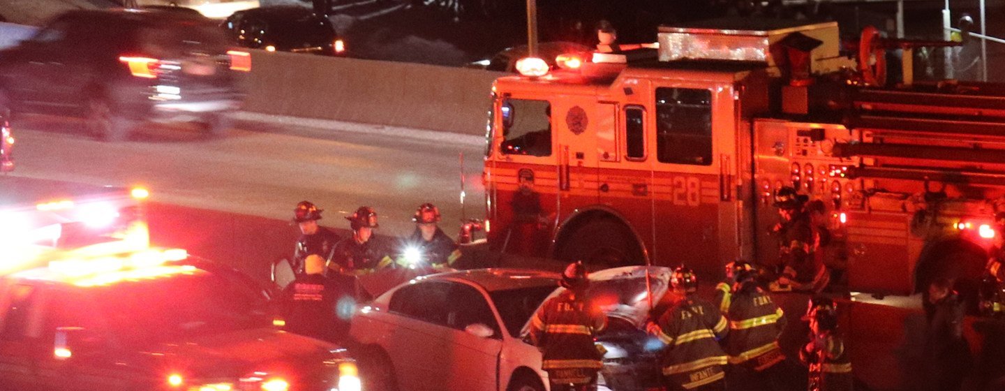 夜幕降临，在高速公路上，一辆白色轿车受损，消防车的红色警示灯闪烁，身穿黄黑相间制服的消防员正在现场进行抢修。