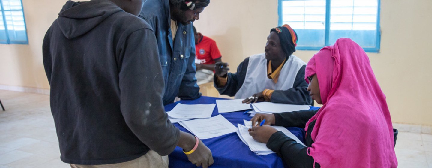 Un groupe de personnes, dont deux hommes et deux femmes, sont réunis autour d'une table recouverte d'une nappe bleue, en train de s'inscrire ou de signer des documents à l'intérieur d'un bâtiment.