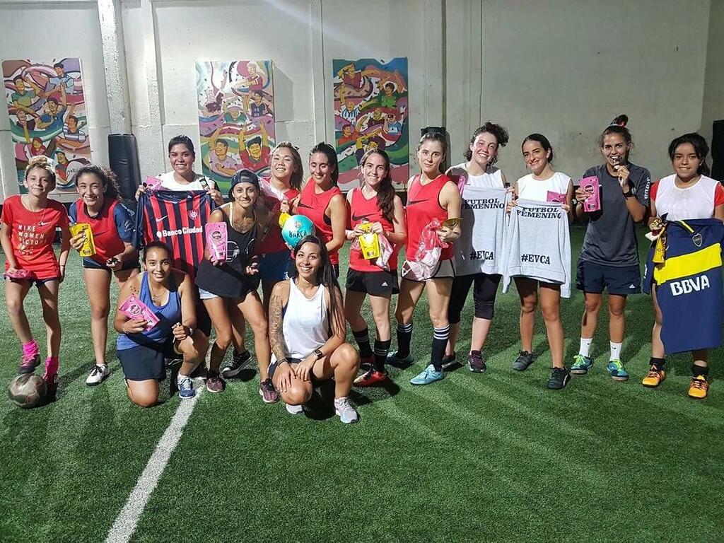 Un grupo de jugadoras de fútbol posa para una foto en una cancha de césped artificial cubierta, sosteniendo camisetas y trofeos.