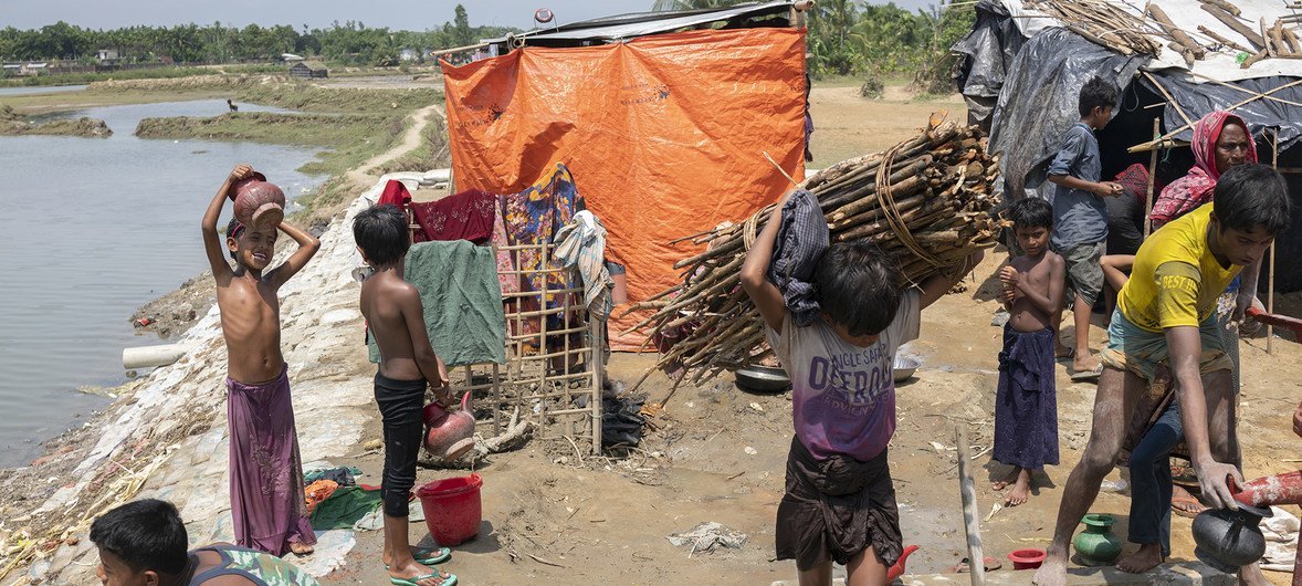 一个罗兴亚男孩扛着一堆柴火，其他难民则在考克斯巴扎难民营内的一口管井边洗澡。这一区域就在涨潮时的水位线上，非常靠近大海，极易受到暴风雨威胁。