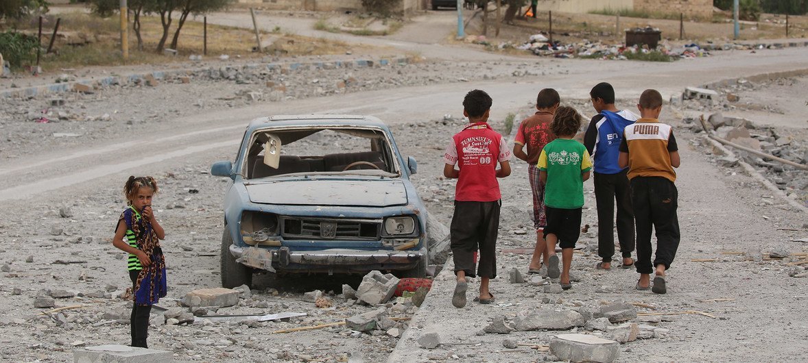穿着红色T恤的12岁叙利亚儿童哈桑（左二）与朋友们走过一辆被炸毁的汽车。哈桑一家已经两次为躲避暴力而逃离家园，使得他被迫从小学三年级辍学。