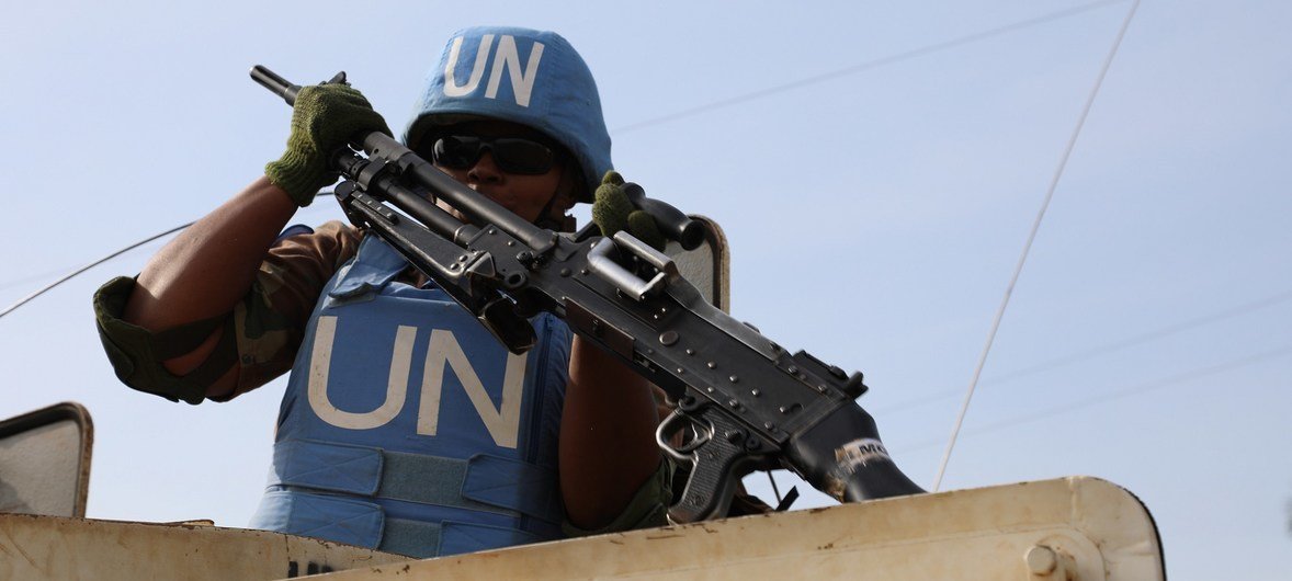 Uma integrante sul-africana das forças de paz da ONU, pertencente à MONUSCO , em patrulha, usando capacete e colete azuis, e segurando um rifle de dentro de um veículo.