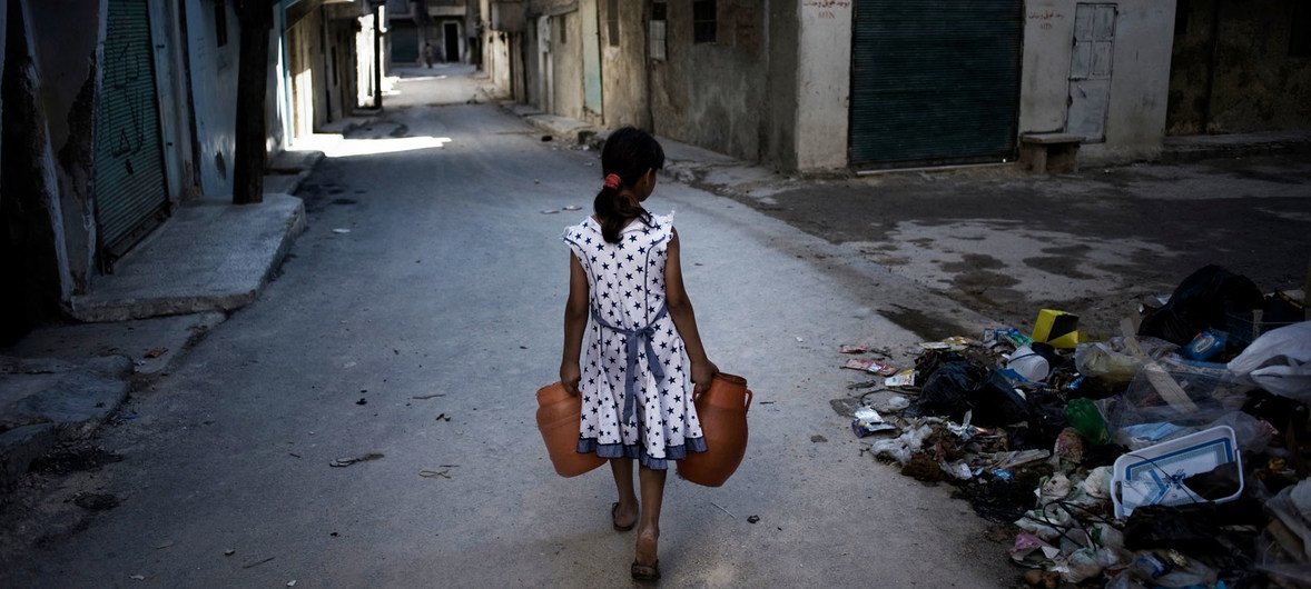  Une fille, transporte des jerrycans d'eau devant une pile de débris dans une rue d'Alep, en Syrie.