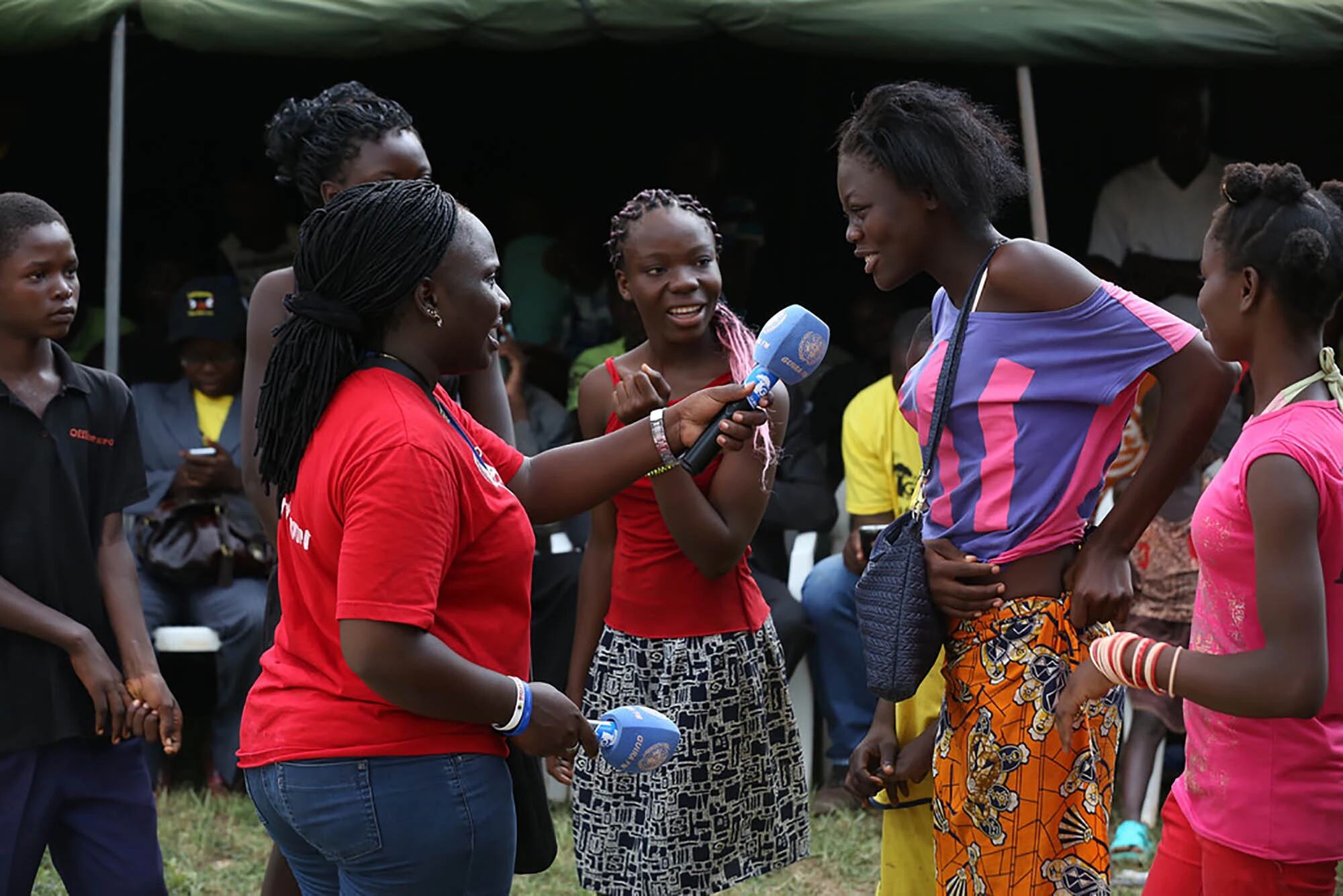 Женщина в красной рубашке интервьюирует молодую женщину с микрофоном на мероприятии на открытом воздухе, организованном радиостанцией ООН Guira-FM, продвигающей мир и примирение в Центральноафриканской Республике.
