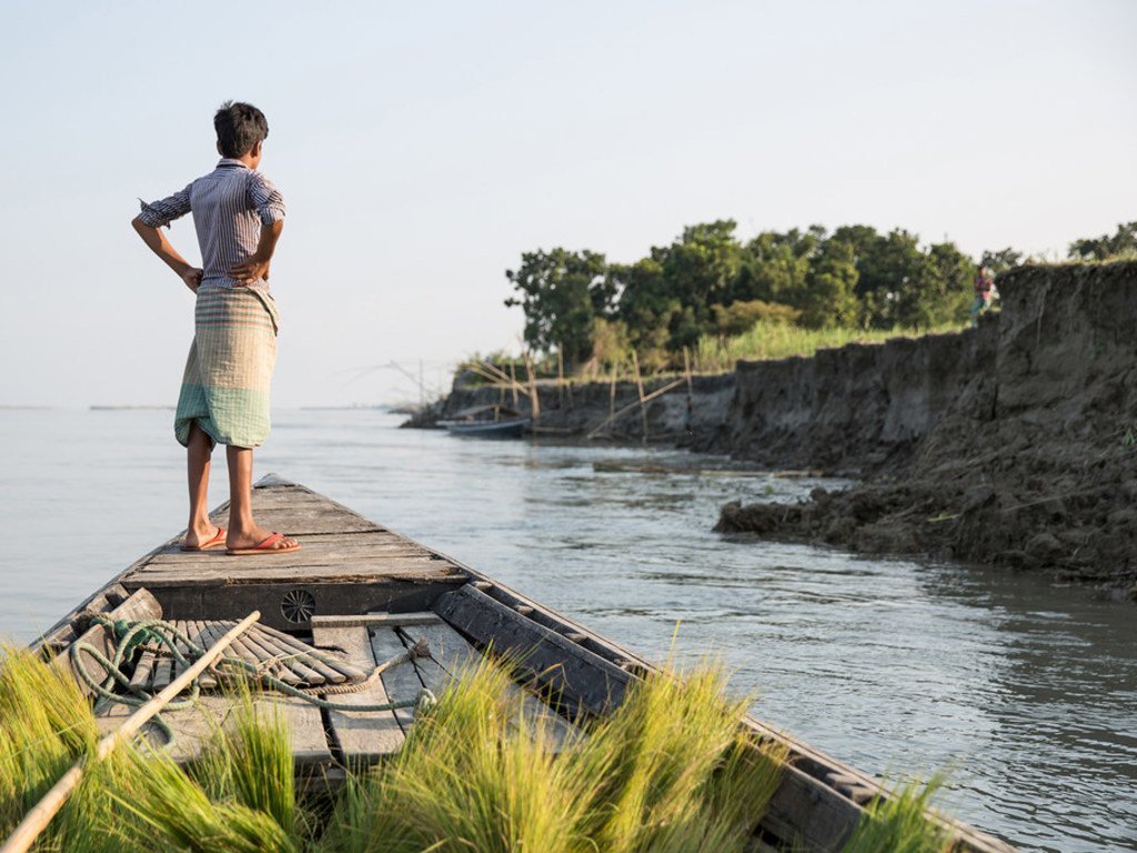 2016年10月，孟加拉国中部锡拉杰甘杰（Sirajganj）县，一个男孩站在船上眺望河岸。由于气候变化，锡拉杰甘杰的农田受到严重侵蚀，许多家庭失去生计，被迫流离失所。