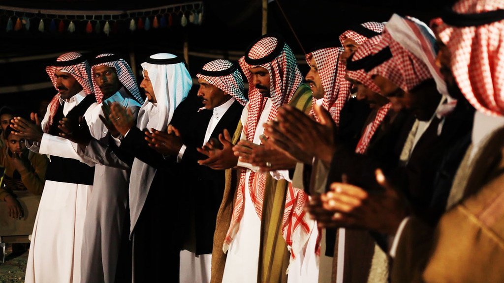 مجموعة من الرجال يرتدون الزي الأردني التقليدي، بما في ذلك الكوفية والثوب، يغنون ويصفقون أثناء عرض فني.