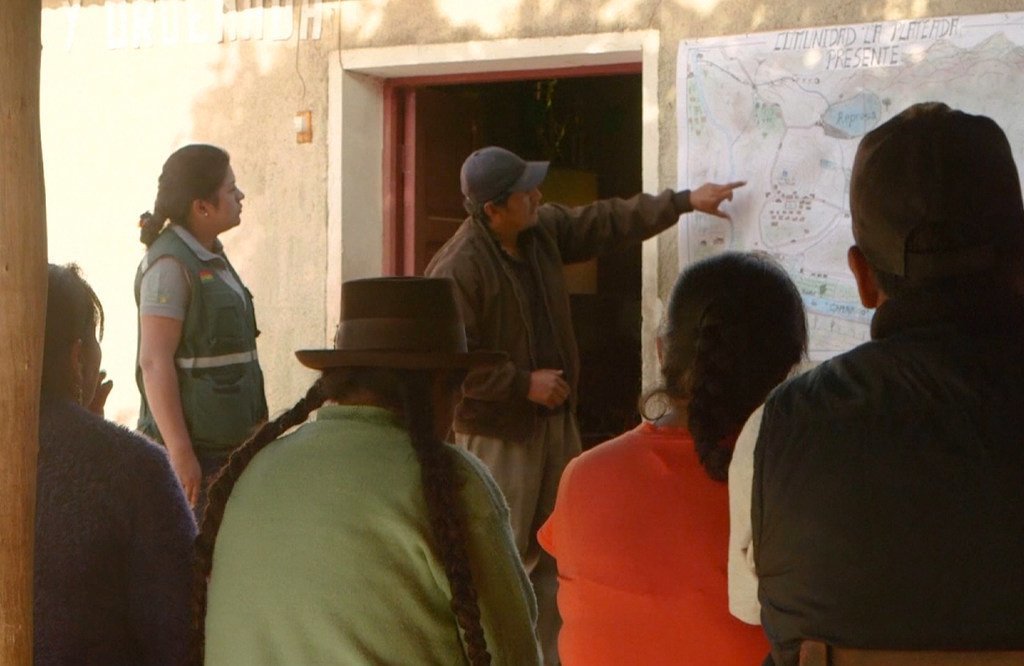 Um homem aponta para um mapa na parede enquanto um grupo de pessoas, incluindo uma mulher com um colete verde e uma pessoa com um chapéu tradicional, observa.
