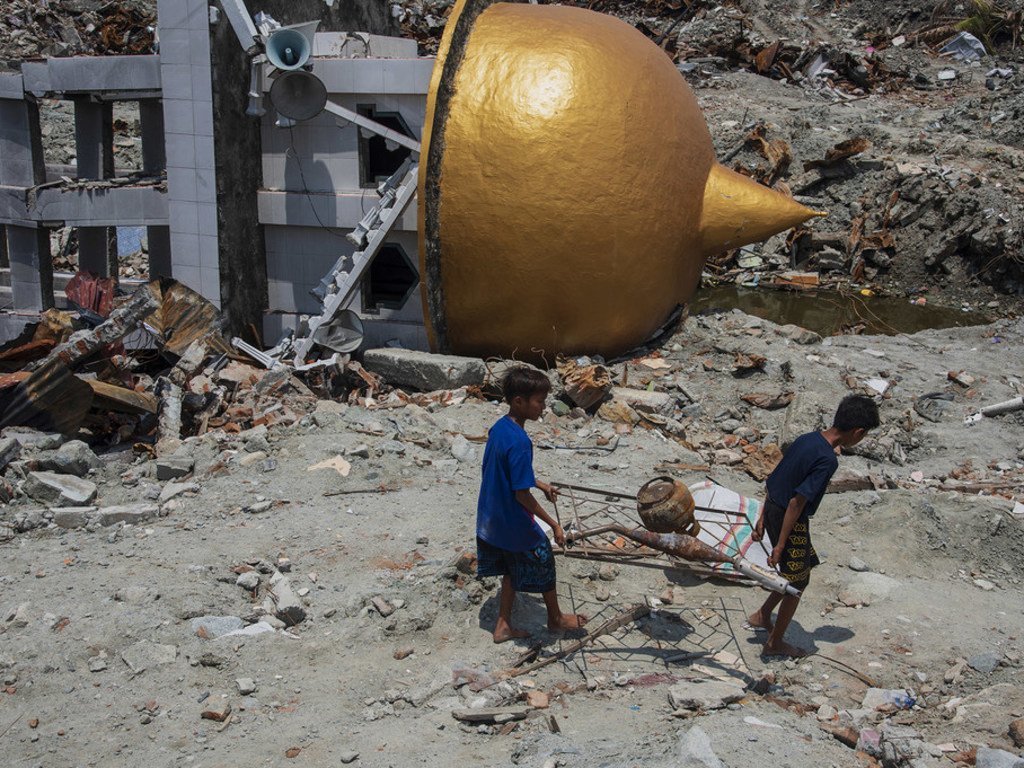 2018年10月17日，在印度尼西亚苏拉威西岛的一处古迹，两个当地居民扛着从废墟中打捞的金属正在从瓦砾堆上和坍塌的清真寺旁走过 。2018年9月28日发生的强烈地震完全摧毁了苏拉威西岛的这处遗址。 
