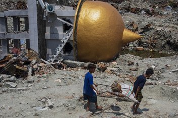2018年10月17日，在印度尼西亚苏拉威西岛的一处古迹，两个当地居民扛着从废墟中打捞的金属正在从瓦砾堆上和坍塌的清真寺旁走过 。2018年9月28日发生的强烈地震完全摧毁了苏拉威西岛的这处遗址。 