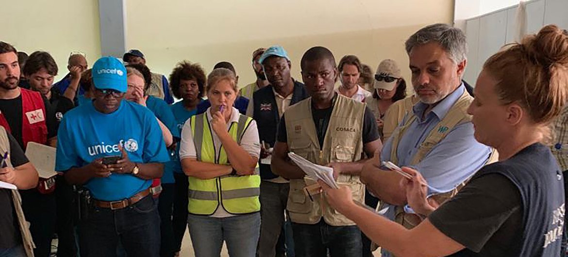 Um grupo de trabalhadores humanitários internacionais e funcionários da ONU , incluindo um homem com uma camisa da Unicef e outros com coletes de segurança, se reúnem em um local fechado em Moçambique para coordenar os esforços de ajuda pós-ciclone.