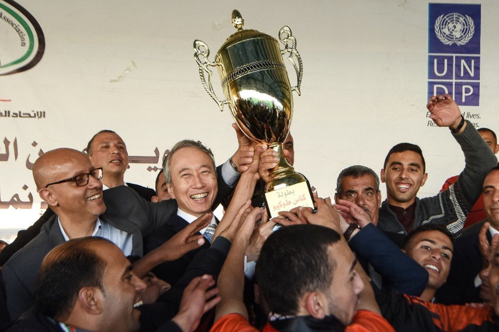 السفير الياباني تاكيشي أوكوبو، وممثل برنامج الأمم المتحدة الإنمائي يحملان كأس بطولة دوري طوكيو النهائية في غزة لتقديمه إلى فريق خان يونس الفائز بالبطولة.