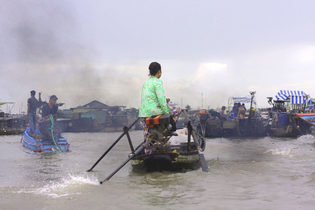 Une Vietnamienne vêtue d'un chemisier vert à motifs manœuvre une pirogue à longue queue sur un fleuve du Vietnam, avec d'autres bateaux et de la fumée visibles en arrière-plan.