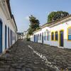 Uma rua de paralelepípedos no centro histórico de Paraty, Brasil, ladeada por prédios coloniais brancos com portas e janelas azuis e amarelas.
