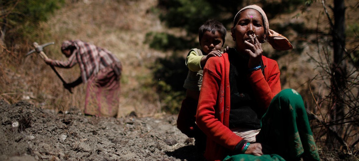 A woman, her child behind her, pauses in the field to smoke a cigarette in Sawa Khola Village, Mugu District.