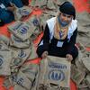 UNHCR کی بنیان پہنے ہوئے ایک عورت نارنجی رنگ کے ٹرپ پر بیٹھی ہے جس کے چاروں طرف UNHCR کے برانڈ والے ٹوٹ بیگز بھرے ہوئے ہیں۔