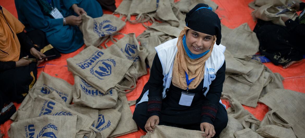 UNHCR کی بنیان پہنے ہوئے ایک عورت نارنجی رنگ کے ٹرپ پر بیٹھی ہے جس کے چاروں طرف UNHCR کے برانڈ والے ٹوٹ بیگز بھرے ہوئے ہیں۔