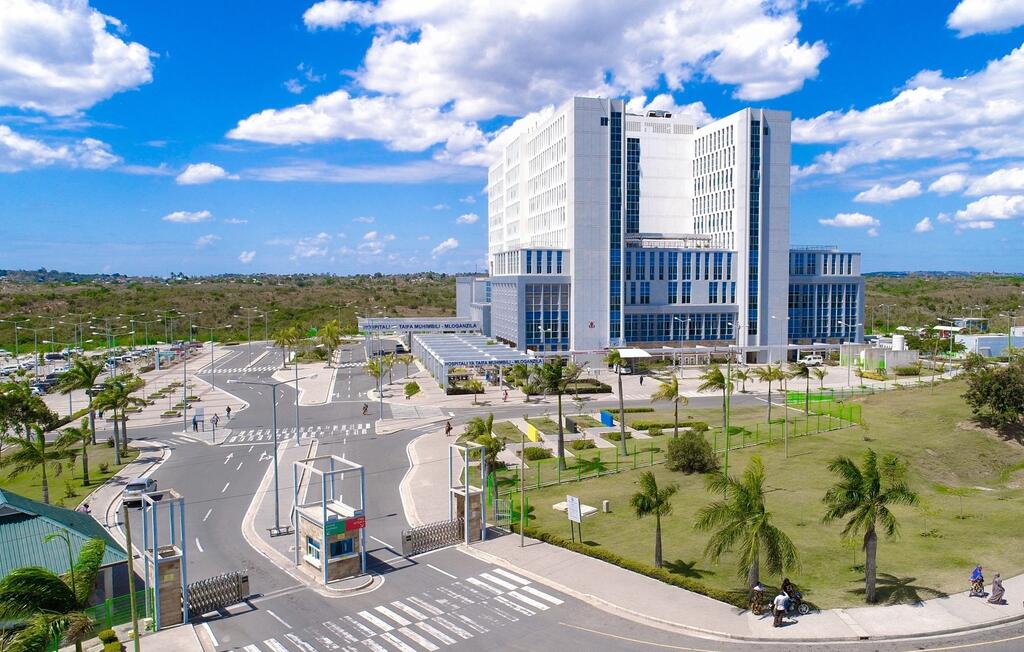 Mtazamo mpana wa Hospitali ya Taifa ya Muhimbili nchini Tanzania, jengo kubwa la kisasa jeupe lenye madirisha ya kioo, lililowekwa dhidi ya anga la bluu lenye mawingu.
