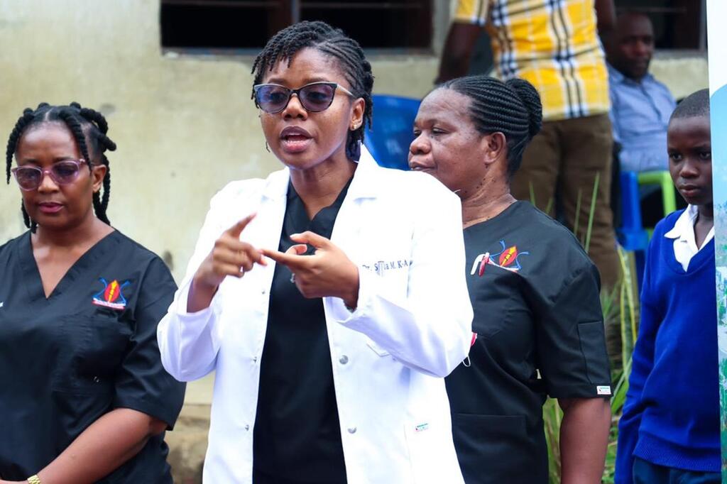 Mtaalamu wa afya wa kike aliyevaa koti jeupe na miwani ya jua akizungumza na kundi la watu katika hafla ya nje nchini Tanzania.