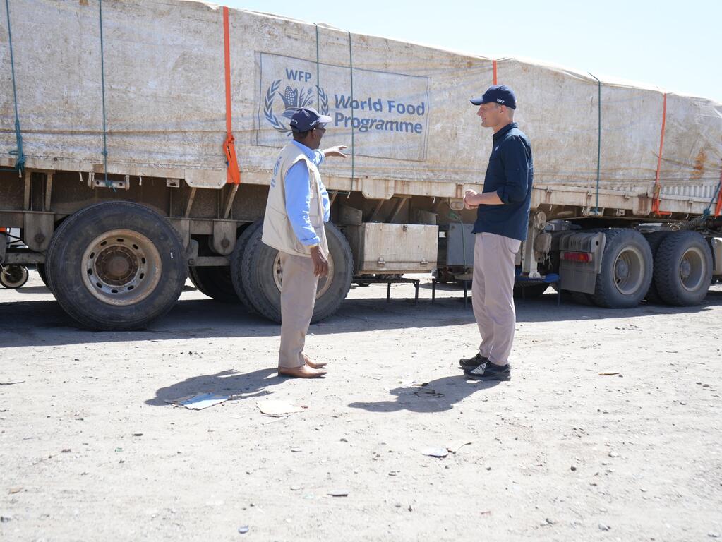 Un travailleur humanitaire du PAM et un autre homme discutent à côté d'un gros camion portant le logo du Programme alimentaire mondial, dans un décor extérieur poussiéreux.