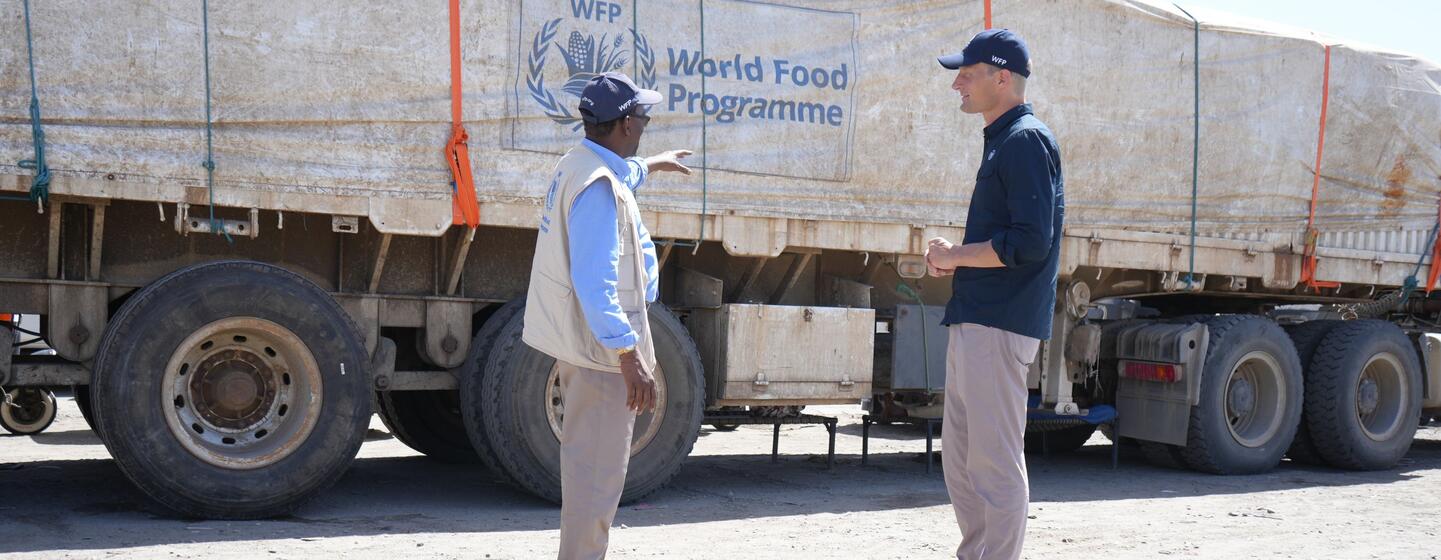 عامل إغاثة من برنامج الأغذية WFP ورجل آخر يتحدثان بجوار شاحنة كبيرة تحمل شعار برنامج الأغذية العالمي في مكان خارجي مغبر.