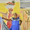 Une femme en robe bleue pose devant une fresque représentant un vendeur de fruits et un boxeur.