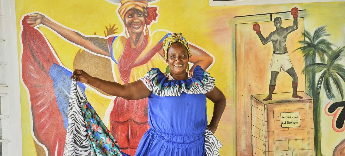 Una mujer con un vestido azul posa frente a un mural que representa a un vendedor de frutas y a un boxeador.